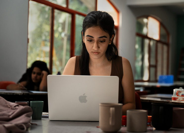 A person working on a Macbook in a classroom, focused on their screen with cups and bagpack on the table.