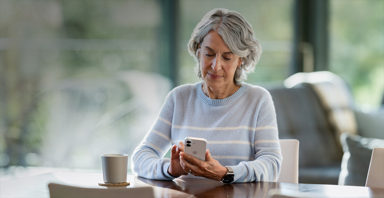 سيدة مسنة جالسة إلى طاولة بينما تنظر إلى جهازها iPhone.‏