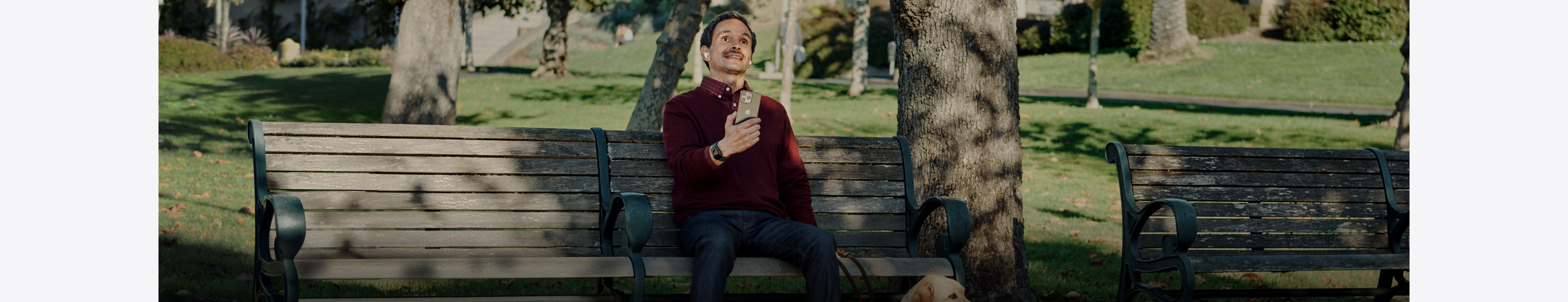 Man with vision disability sitting outside on a park bench while using vision accessibility feature on iPhone and wearing AirPods.