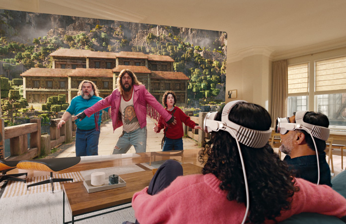 Zwei Personen, die beide jeweils eine Apple Vision Pro tragen, sitzen nebeneinander auf der Couch und schauen: Ein Minecraft Film, gezeigt auf einem Bildschirm, der sich durch den gesamten Raum erstreckt